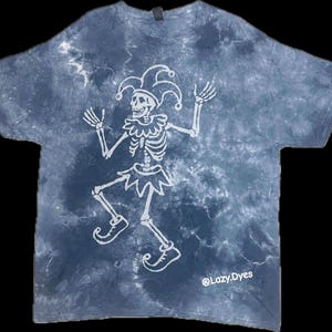 May include: A tie-dye t-shirt in shades of blue and white, featuring a white skeleton jester design. The jester is wearing a hat, collar, and skirt. The text "@Lazy.Dyes" is printed on the bottom right of the shirt.