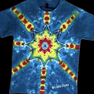 Op de afbeelding: Blauw tie-dye T-shirt met een centraal sterpatroon in geel, rood en wit. Het shirt heeft een ronde hals en korte mouwen. De tekst "@Lazy.Dyes" staat onderaan. Het tie-dye patroon straalt uit het midden.