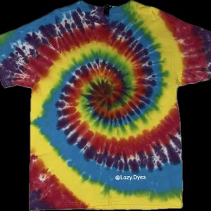 May include: A short-sleeved t-shirt with a spiral tie-dye pattern in shades of yellow, red, blue, green, and purple. The shirt has a crew neck and the text "@Lazy.Dyes" is visible.