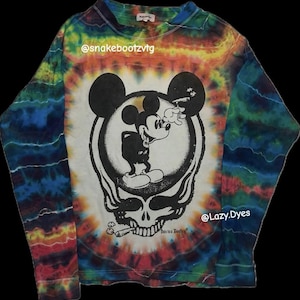 Puede incluir: Camiseta de manga larga teñida con un diseño de Mickey Mouse fumando un cigarrillo, superpuesto a un diseño de calavera. La camiseta tiene un esquema de color arcoíris y el texto "this is a Bootleg" está impreso en la parte inferior.