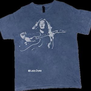 Puede incluir: Camiseta azul con un gráfico blanco de un músico tocando la guitarra. La ilustración es una imagen estilizada. El texto "@Lazy.Dyes" está impreso en la parte inferior de la camiseta.