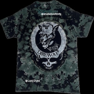 Puede incluir: Camiseta teñida con tinte anudado en tonos verdes y negros. Presenta un diseño de lobo con pañuelo, las palabras "Dire Wolf" y un diseño de calavera. También se ven los textos "@snakebootzvtg" y "@Lazy.Dyes".