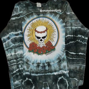 Puede incluir: Camiseta de manga larga teñida con un diseño central de una calavera con una corona floral, rodeada de un sol y rosas. La camiseta tiene un patrón de teñido anudado azul, blanco y negro. El texto en la camiseta dice "@snakebootzvtg" y "@Lazy.Dyes".
