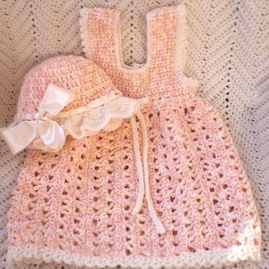 May include: A pink crocheted baby dress with a matching crocheted hat and a white satin bow. The dress has a scalloped edge and a white crocheted trim.