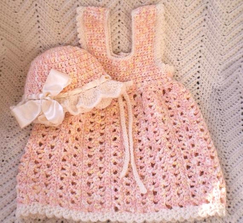May include: A pink crocheted baby dress with a matching crocheted hat and a white satin bow. The dress has a scalloped edge and a white crocheted trim.