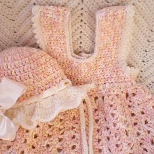 May include: A pink and yellow crocheted baby dress and matching hat with a white satin bow. The dress and hat are made with a delicate crochet stitch.