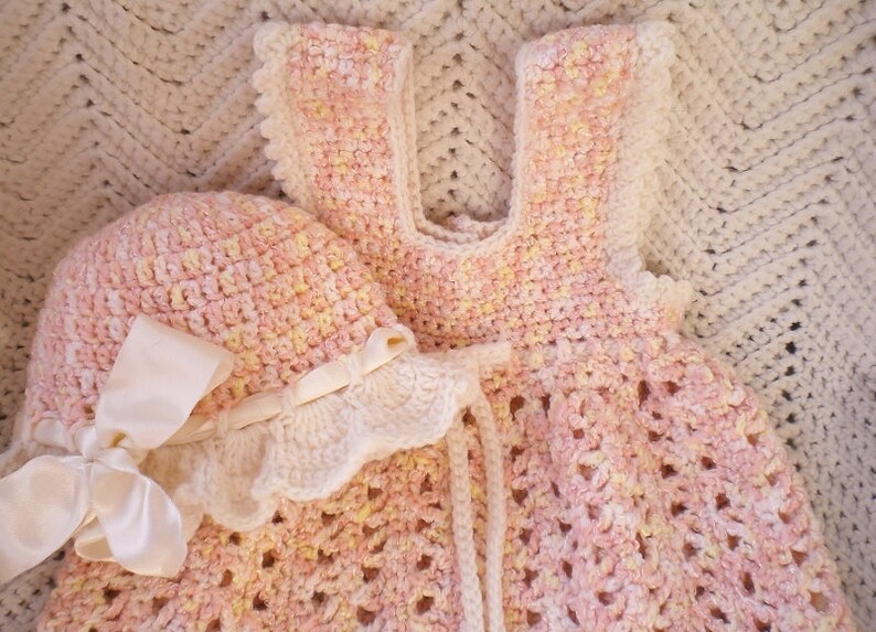 May include: A pink and yellow crocheted baby dress and matching hat with a white satin bow. The dress and hat are made with a delicate crochet stitch.