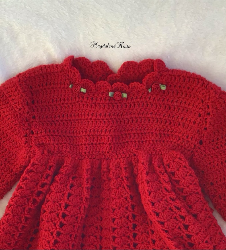 May include: A red crocheted baby dress with a scalloped neckline and green ribbon embellishments. The dress has a simple design and is made with a soft, textured yarn. The text 'Magdalene Knits' is visible in the top center of the image.