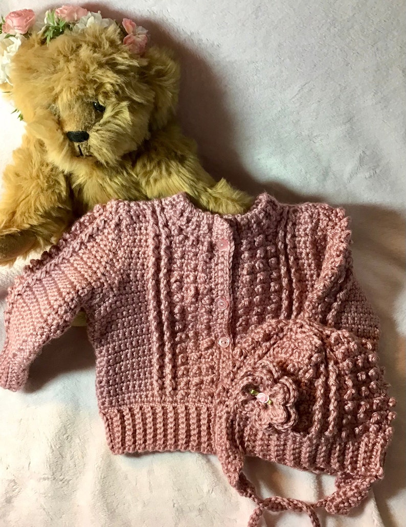 May include: A pink crocheted baby cardigan and matching hat with a flower embellishment