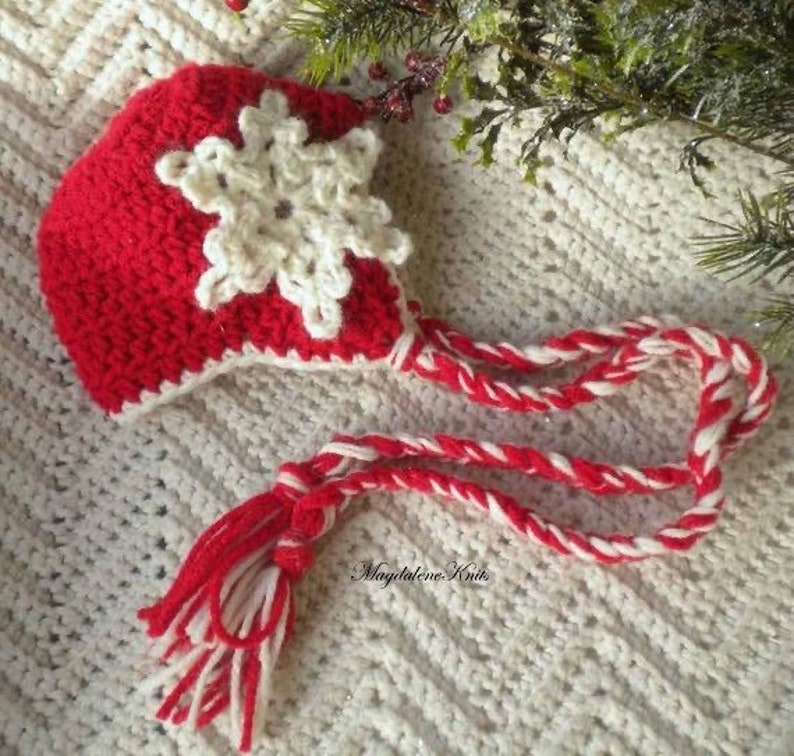 May include: A red and white crocheted hat with a white snowflake design. The hat has a long, red and white braided cord with a tassel at the end. The text 'Magdalene Knits' is visible on the cord.