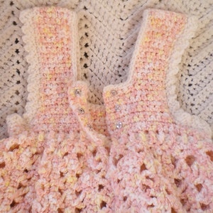 May include: A crocheted baby dress in pink and yellow with a scalloped edge and button closures.