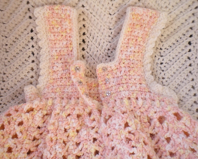May include: A crocheted baby dress in pink and yellow with a scalloped edge and button closures.