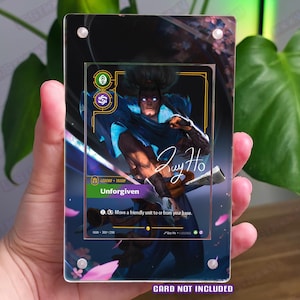 May include: A rectangular acrylic display case featuring a printed card with a character illustration, the text Unforgiven, Legend Yasuo, and a signature. The case has four silver corner screws and a dark background with pink cherry blossom petals.