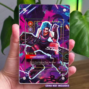 Pode incluir: Um suporte de cartão acrílico transparente com uma ilustração colorida de um personagem com cabelo azul e o texto "Loose Cannon". O suporte do cartão tem um design vibrante com detalhes em roxo, rosa e azul. O texto "CARD NOT INCLUDED" está na parte inferior.