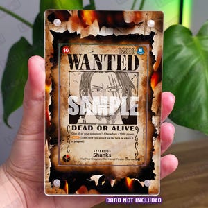 Può includere: Un espositore rettangolare in acrilico con design a bordi bruciati e motivi a fiamma arancioni e neri. Contiene una carta collezionabile One Piece Shanks Wanted con il testo WANTED, 12000 e DEAD OR ALIVE. È presente la scritta CARD NOT INCLUDED.