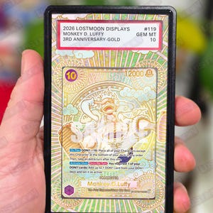 May include: A collectible trading card featuring Monkey D. Luffy from the 2026 Lostmoon Displays series. The card has a gold and white design with the text "3rd Anniversary-Gold" and "GEM MT 10". The card is held in a black protective case.