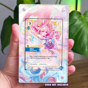 May include: A clear acrylic card holder displaying a colorful card featuring a pink-haired anime character named Ahri. The card has a gold border and text that reads "Ahri INQUISITIVE". The holder is held in a hand.