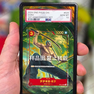May include: A graded trading card featuring Zoro from the 2024 One Piece 1st Anniversary Set. The card displays a colorful illustration of Zoro wielding swords, with Japanese text and a Gem Mint 10 grade.