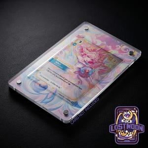May include: A clear acrylic magnetic display case holding a trading card featuring an anime-style character named Ahri. The card has pink, blue, and white swirling background art with text detailing game mechanics and the phrase Just remember to follow the fox.