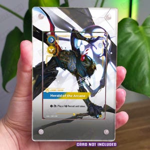 Puede incluir: Una vitrina acrílica transparente que contiene una tarjeta coleccionable. La tarjeta presenta una ilustración de fantasía de una criatura con el texto "Herald of the Arcane". El texto "CARD NOT INCLUDED" está en la parte inferior. La vitrina está delante de una planta verde.