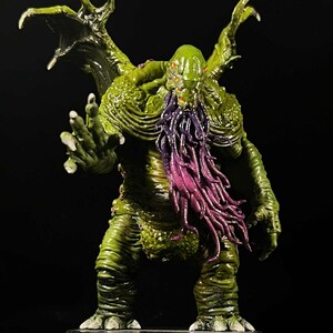 May include: A detailed miniature figure of a monstrous creature with a green, textured body, large wings, and a purple, tentacled beard. The figure has multiple red eyes and is posed on a black base, set against a dark background.