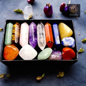 May include: A black box containing an assortment of colourful crystals and gemstones. The crystals are in various shapes, including pointed wands and rounded stones. Colours include green, yellow, orange, pink, purple, clear, and blue. The box has the text "AVA LUXE ILLUMINATIONS".