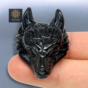Puede incluir: Una figura de cabeza de lobo negra tallada. Los rasgos del lobo son detallados, con textura de pelaje visible y una expresión concentrada. La figura se sostiene en una mano, con el logotipo de AVA LUXE ILLUMINATIONS visible.