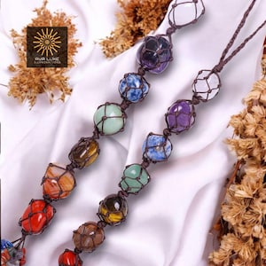 May include: Two necklaces featuring a variety of coloured gemstones, each held within a brown woven net. The gemstones are in shades of red, orange, yellow, green, blue, purple, and white. The necklaces are presented against a white fabric backdrop.