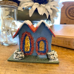Puede incluir: Una casa azul en miniatura con un tejado rojo y detalles de nieve blanca. Las ventanas están delineadas con cuentas rojas y azules, y la casa se asienta sobre una base verde con arbustos nevados. Un tarro con luces de hadas está en el fondo.