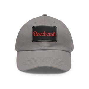 May include: A gray baseball cap with a curved brim. The cap features a black rectangular patch with the word "Beechcraft" in red lettering.