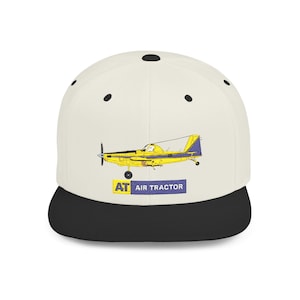 May include: A white and black baseball cap featuring a yellow and blue airplane graphic. The graphic includes the text "AT AIR TRACTOR" in a blue rectangle. The cap has a flat brim and a snapback closure.