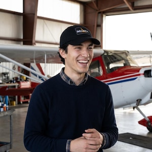 May include: A black baseball cap featuring a rectangular patch with the text BELLANCA in white letters and a stylized yellow wing graphic. The cap has a curved brim and a structured crown, worn by an individual in a dark blue sweater and plaid shirt.