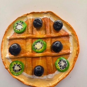 May include: A round, painted artwork resembling a waffle, adorned with blueberries and kiwi slices. The waffle is a warm orange-brown color, with a white border. The kiwi slices are green with black seeds, and the blueberries are dark blue.
