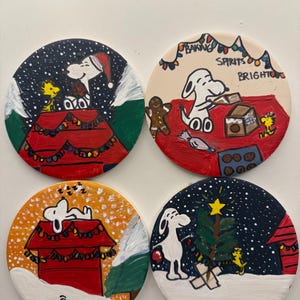 May include: Set of four round coasters featuring Snoopy and Woodstock in holiday scenes. Each coaster has a different design, including Snoopy in a Santa hat, baking, sleeping, and decorating a Christmas tree. The coasters are hand-painted with a white base and colorful details.