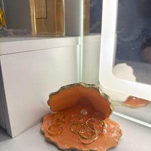May include: An orange, seashell-shaped jewelry dish with gold trim, holding several gold rings and a pearl. The dish sits on a white surface, with a perfume bottle in the background.