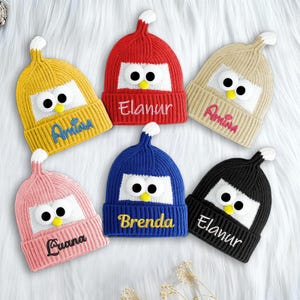 Op de afbeelding: Een verzameling van zes gebreide mutsen met pinguïnthema in verschillende kleuren: geel, rood, beige, roze, blauw en zwart. Elke muts heeft een wit gezicht met grote zwarte ogen en een gele snavel, en een geborduurde naam.