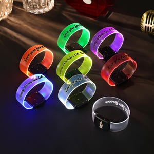 Może przedstawiać: Kolekcja podświetlanych opasek na nadgarstek w różnych kolorach, w tym pomarańczowym, zielonym i fioletowym. Każda opaska zawiera spersonalizowany tekst, taki jak "Love you forever" oraz imiona z datami. Opaski mają czarną, prostokątną podstawę.