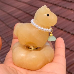 May include: A carved, light yellow jade horse figurine with a pearl necklace and a small green jade pendant. The horse is mounted on a round base with a gold-coloured metal accent. The figurine is held in a person's hand.