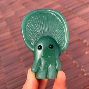 May include: A green mushroom-shaped figurine with a smiling face and black eyes. The figurine has a detailed cap with vertical lines and a smooth, rounded body. The figurine is held in a hand.