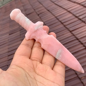 Puede incluir: Una daga de cristal rosa con un mango redondeado y una hoja puntiaguda. La daga tiene un diseño tallado y un color rosa claro con algunas vetas blancas y grises. La daga se sostiene en una mano.