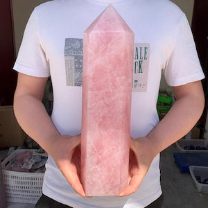 May include: A large, pink rose quartz crystal point. The crystal is smooth and polished, with a pointed tip and a flat base. The crystal is being held by a person's hands.