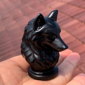 May include: A glossy black wolf head sculpture. The wolf's head is carved with detailed fur and features, mounted on a circular base. The sculpture is held in a hand against a blurred background.