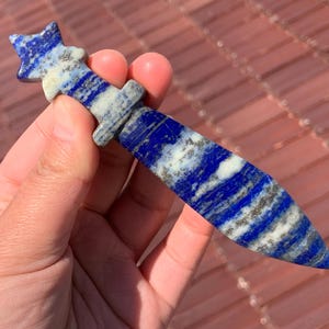 May include: A blue and white lapis lazuli sword shaped crystal. The crystal is polished and has a smooth surface.