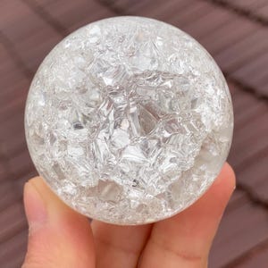 May include: A clear, spherical crystal ball with a cracked, textured surface. The ball is held up against a brown background. The sphere is a decorative item.