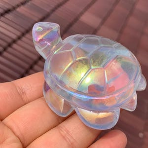 Puede incluir: Figura de tortuga de cristal iridiscente con acabado pulido. La figura tiene un patrón de caparazón detallado y patas, cabeza y cola visibles. El cristal refleja un arcoíris de colores, incluyendo rosa, amarillo y azul. La tortuga es un objeto decorativo.