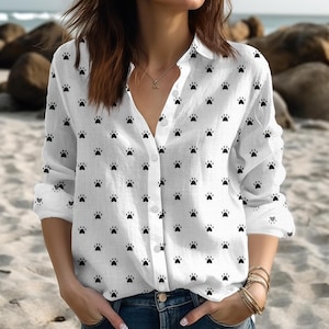 May include: White button-down shirt with a repeating pattern of black paw prints. The long-sleeved shirt has a collared neckline and is paired with blue jeans. The shirt is made of a lightweight fabric.