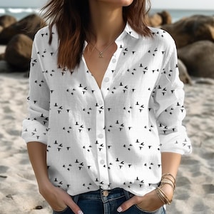 Può includere: Camicia bianca con bottoni, maniche lunghe e motivo a uccelli. La camicia presenta un colletto classico e uno scollo a V. Le sagome nere degli uccelli sono uniformemente distanziate sul tessuto, creando un design semplice ma elegante. La camicia è abbinata a jeans blu.