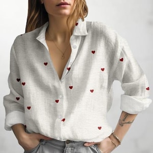 Peut inclure: Chemise grise clair boutonnée ornée de petites broderies de cœurs rouges. La chemise à manches longues a un col et des poignets classiques. La chemise est en tissu texturé.