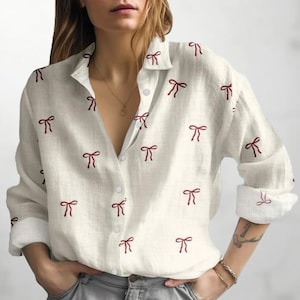 Könnte beinhalten: Ein cremefarbenes Button-Down-Hemd mit langen Ärmeln und einem Muster aus gestickten weinroten Schleifen. Das Hemd hat einen klassischen Kragen und Manschetten und ist aus leichtem Stoff gefertigt.