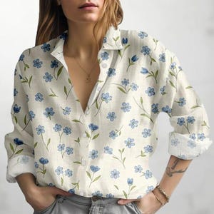 May include: An off-white button-down shirt featuring a floral design. The shirt is patterned with blue flowers and green leaves. It has a collared neckline and long sleeves.